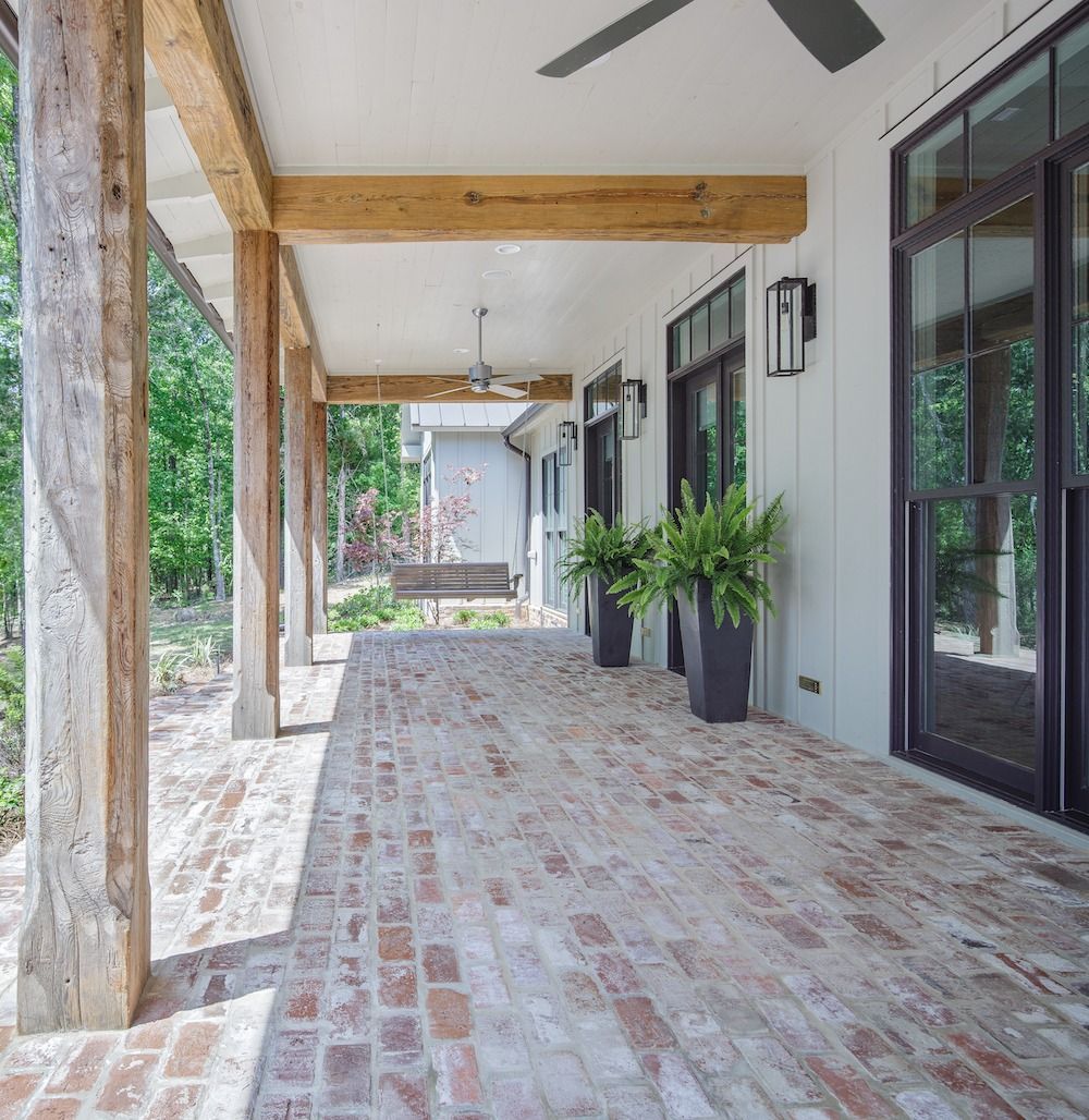 A covered outdoor porch with rustic wood columns, a light brick floor, potted green plants, and dark framed doors.