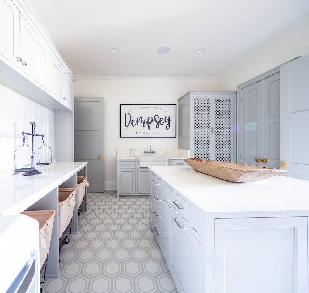 A bright, modern laundry room with pale blue cabinets, quartz countertops, a farmhouse sink, and gray hexagon tile floors.