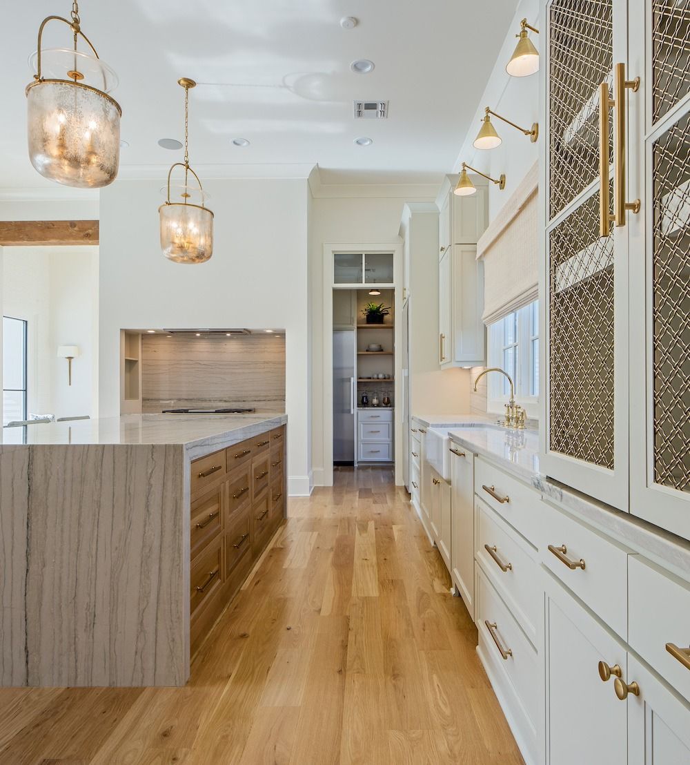 A kitchen with a wood island, two pendant lights, white cabinets, and gold hardware, opening to a pantry.