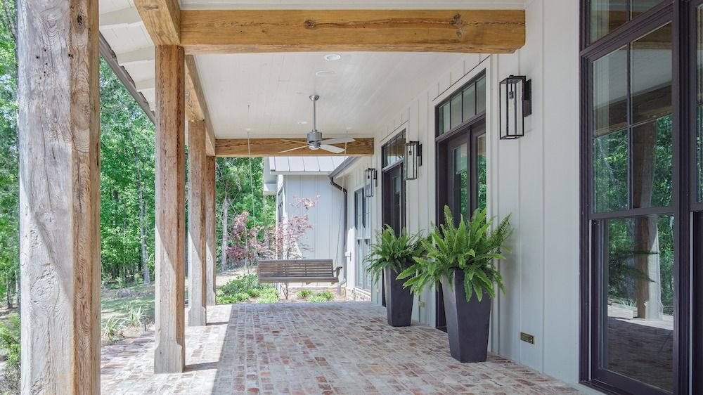 A covered porch with rustic wooden beams, a brick floor, ceiling fan, and potted ferns overlooking a wooded yard.