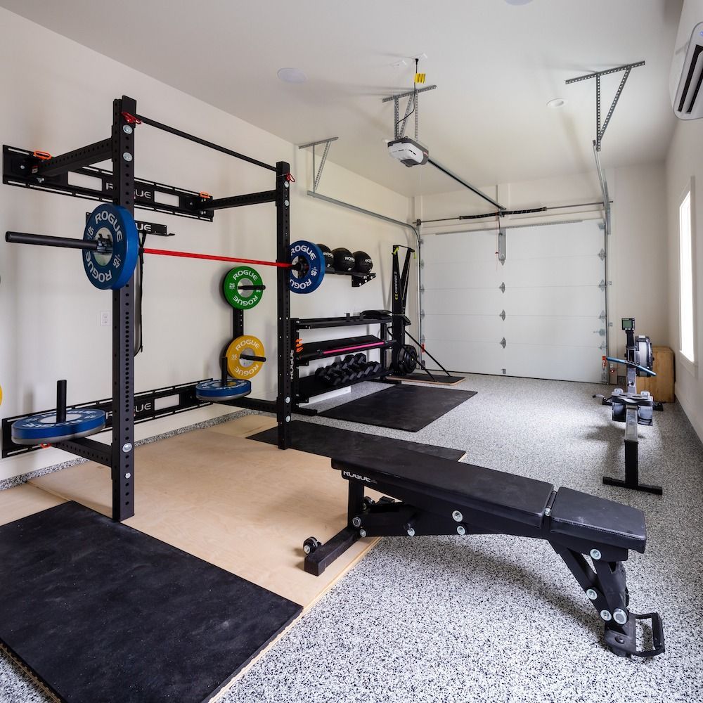 A home garage gym with a wall-mounted rack, barbell, weight plates, an adjustable bench, and a rowing machine.