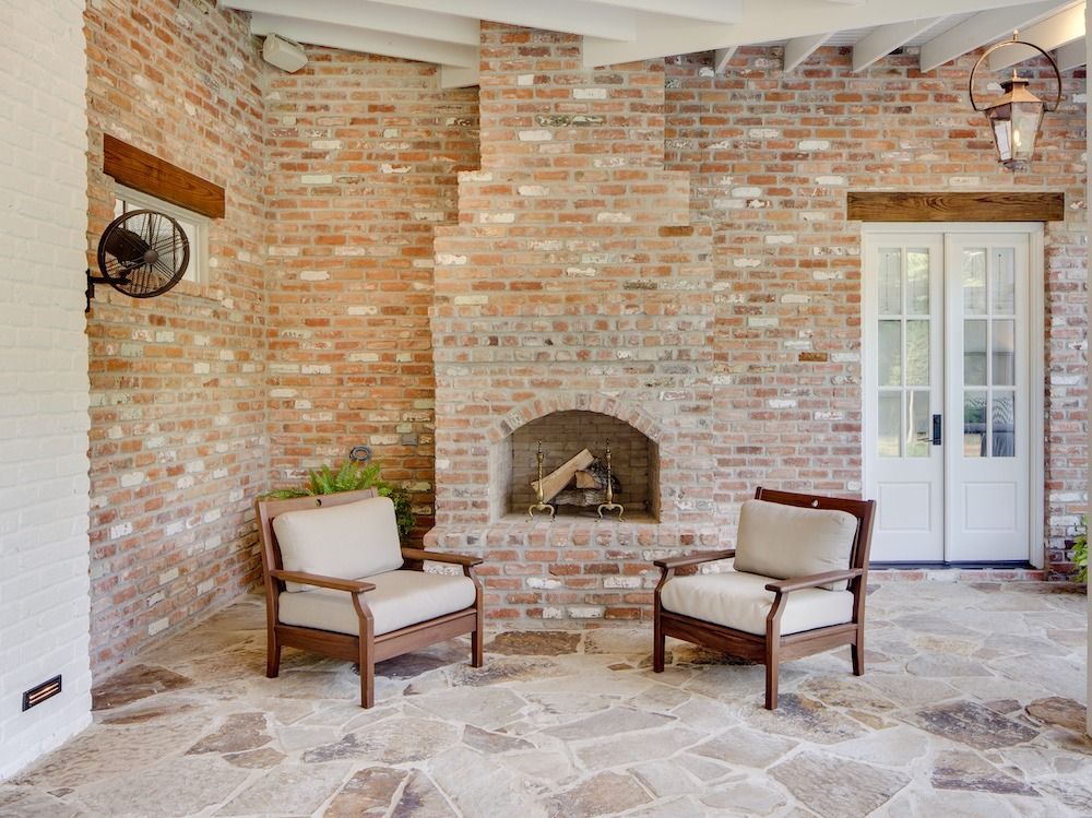 Two armchairs with light cushions face a brick fireplace on a flagstone patio under a white-beamed ceiling.