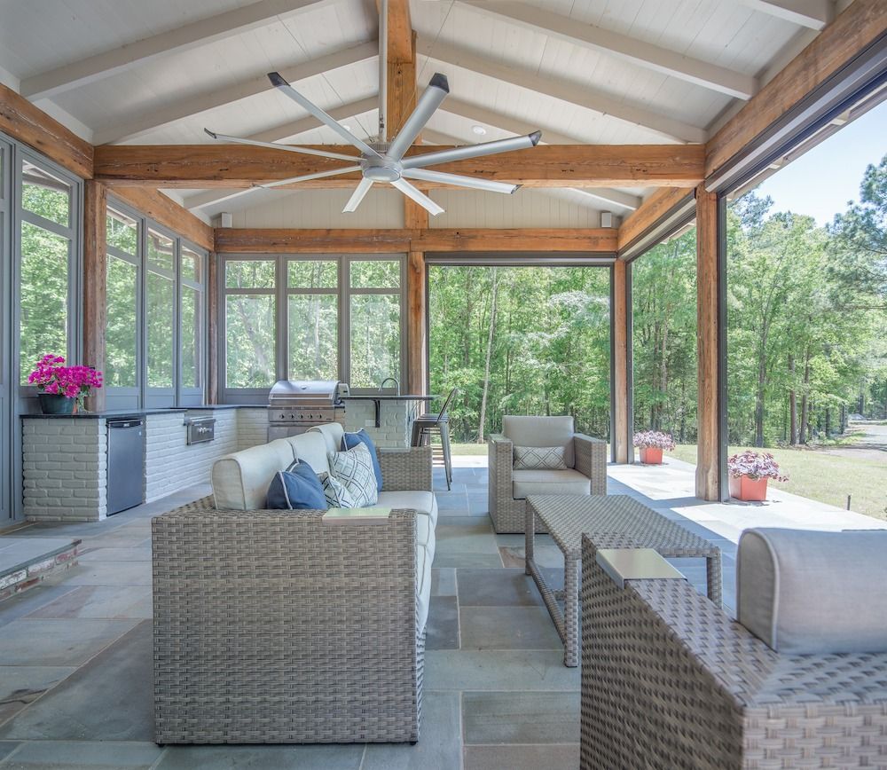 A covered patio with a vaulted ceiling, wicker furniture, and large windows overlooking a wooded outdoor area.