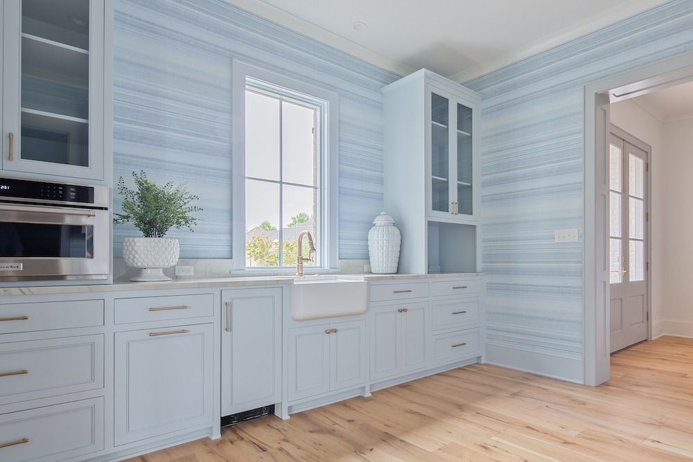 A bright kitchen with light blue cabinetry, striped wallpaper, a farmhouse sink, and hardwood floors.