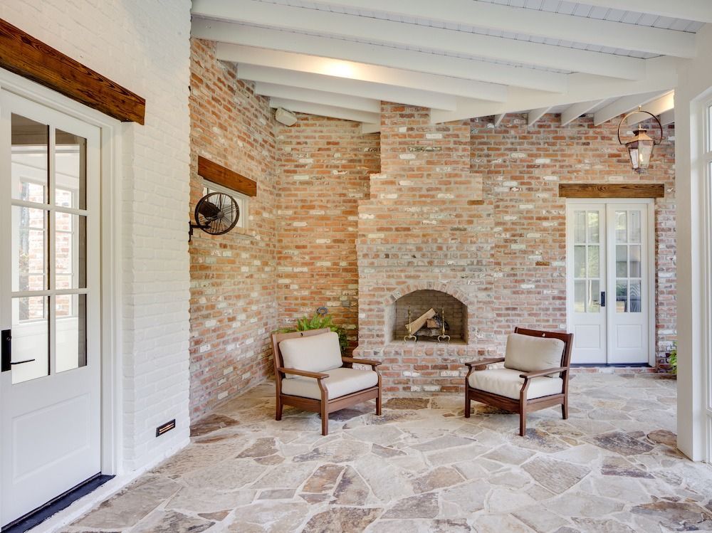 Two armchairs face a brick fireplace on a stone patio with white French doors and a vaulted wood-beam ceiling.