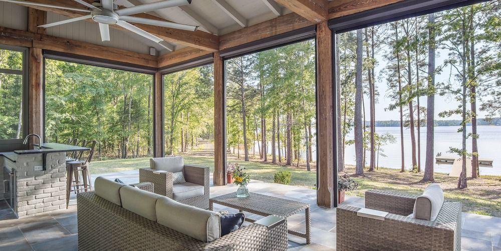 A covered outdoor patio with wicker furniture, a bar, and a ceiling fan, overlooking a lake surrounded by forest.