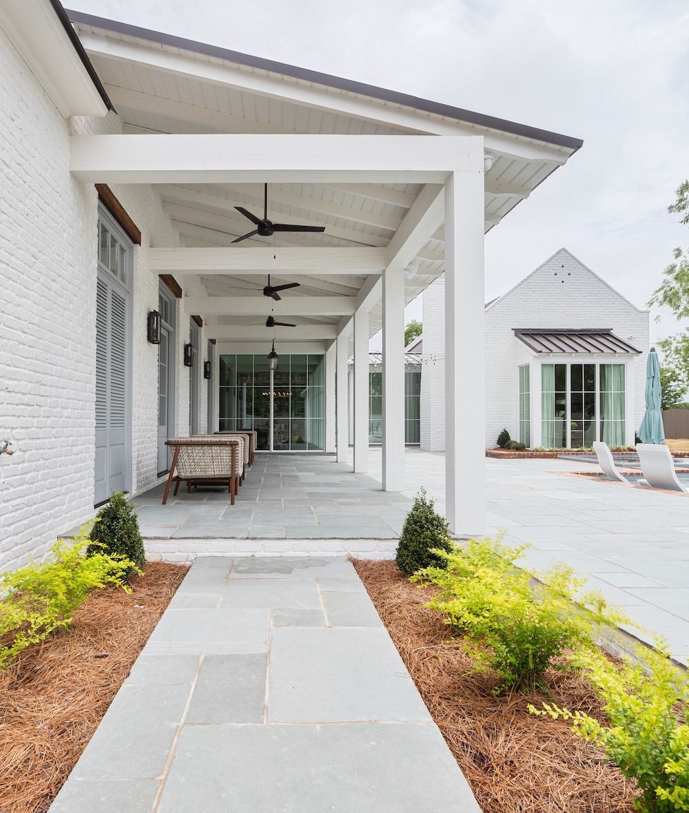 A stone path leads to a white porch with ceiling fans, adjacent to a patio with white chairs and a matching building.