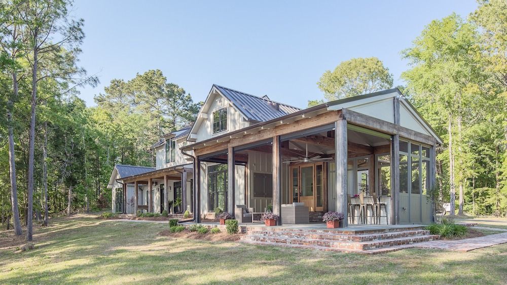 A white farmhouse with a large covered porch and brick patio, nestled in a wooded area on a sunny day.