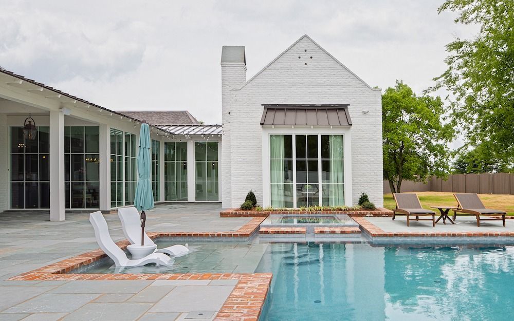 A white home with a backyard pool, stone patio, lounge chairs, and an umbrella on a cloudy day.