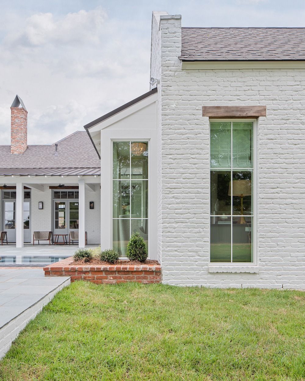 A side view of a white brick house featuring a tall window, a brick flower bed, and a covered patio in the background.