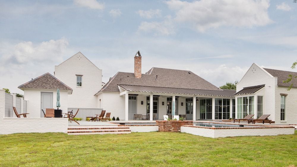 A white cottage-style house with a gray roof and a brick patio sits on a grassy lawn under a partly cloudy blue sky.