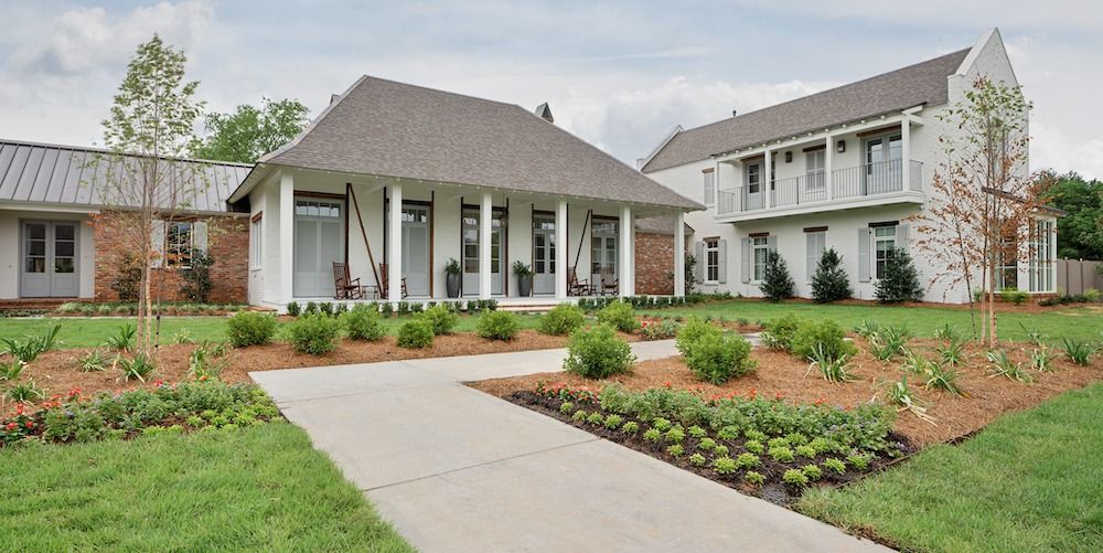 A bright, modern house with white siding and gray shingles, fronted by a sidewalk, manicured lawn, and small shrubbery.