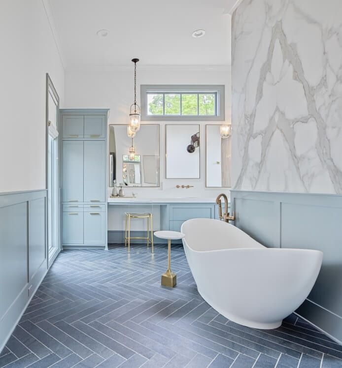 A modern bathroom with blue wainscoting, a freestanding white tub, grey herringbone tile floors, and a marble wall.