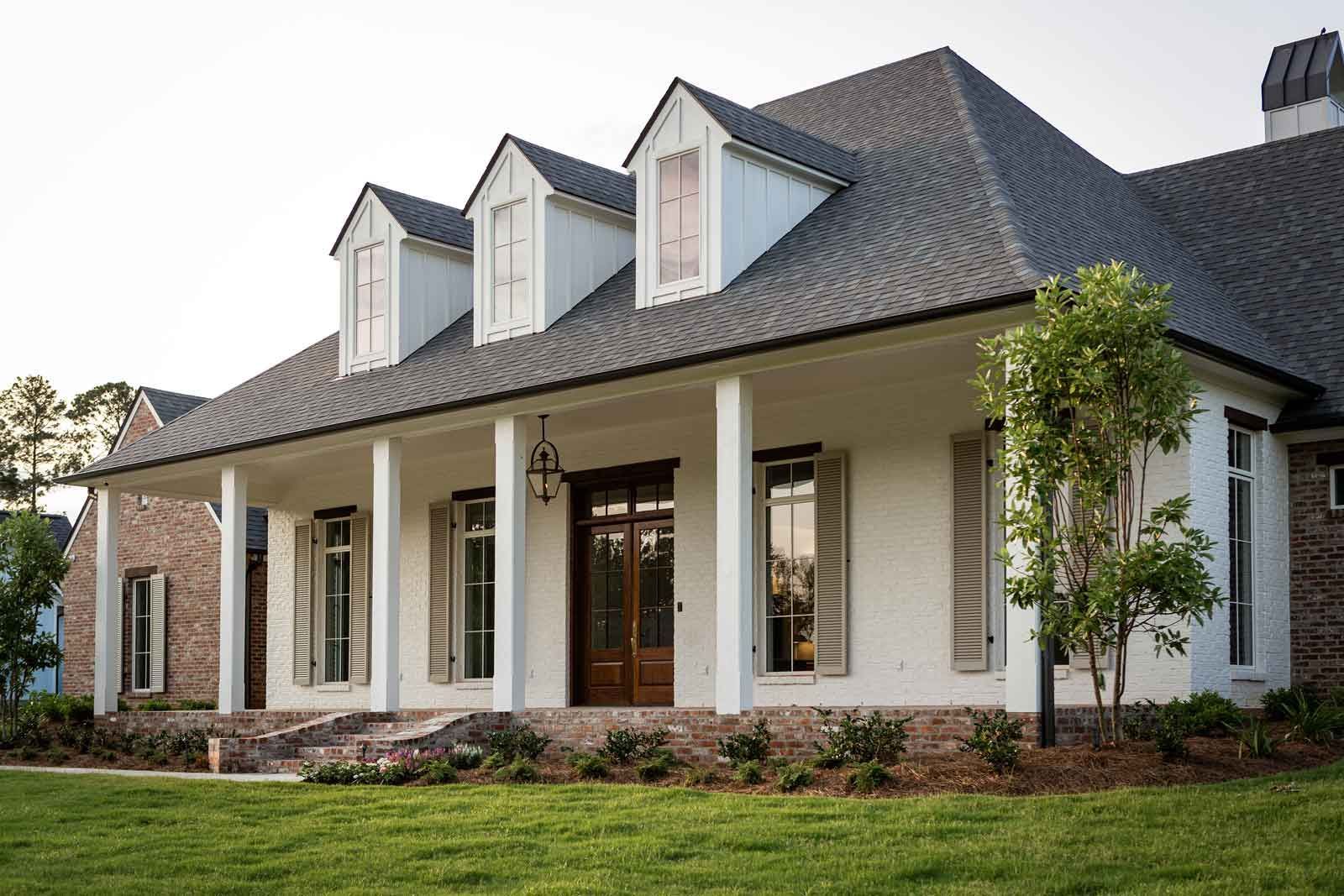 A white, single-story house with a prominent front porch, three roof dormers, dark shutters, and a dark shingled roof.