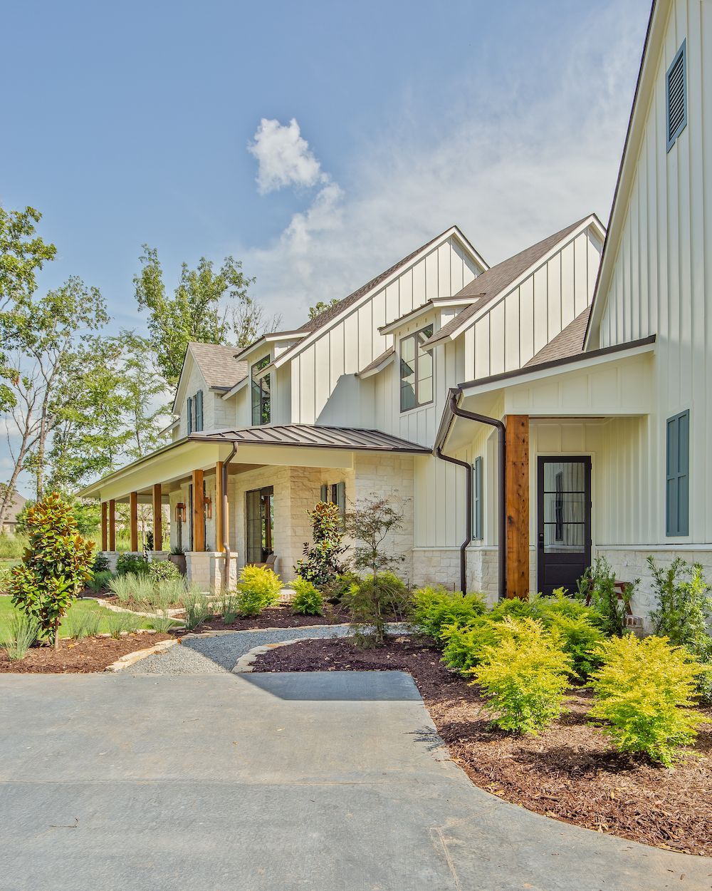 A cream-colored farmhouse with vertical siding, a stone facade, a dark metal roof, and a front porch, surrounded by greenery.