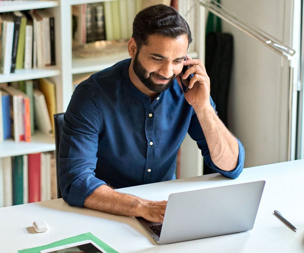 A man is sitting at a desk using a laptop and talking on a cell phone.