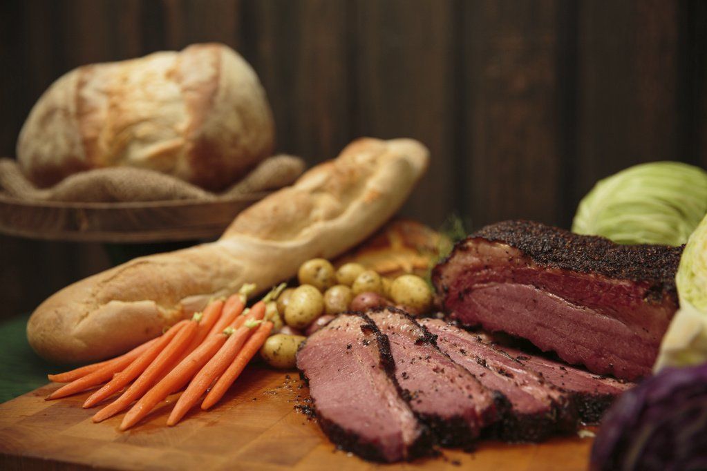 sliced corned beef, loaves of bread, carrots, and potatoes on a wood board.