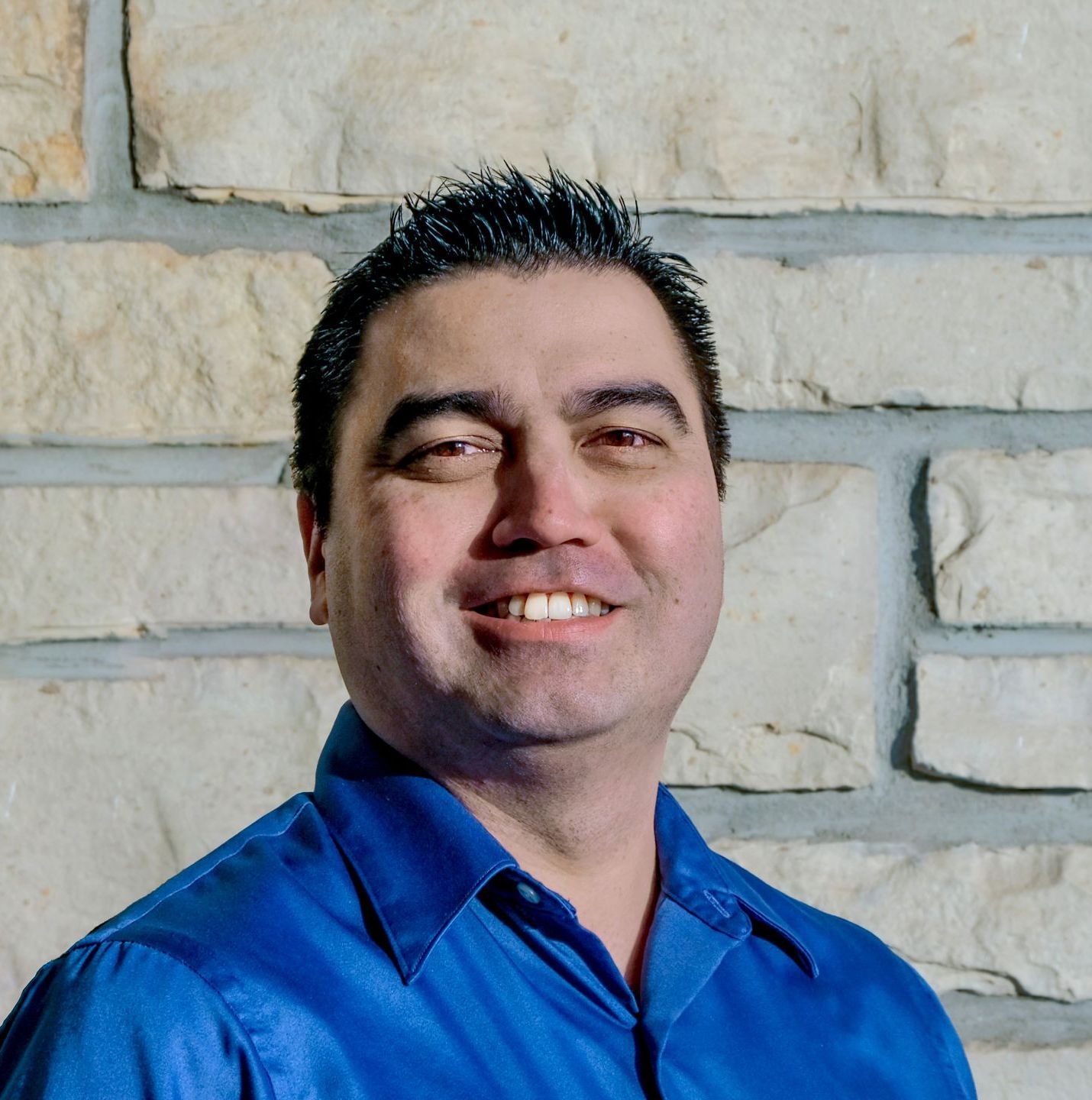 A man in a blue shirt is smiling in front of a brick wall