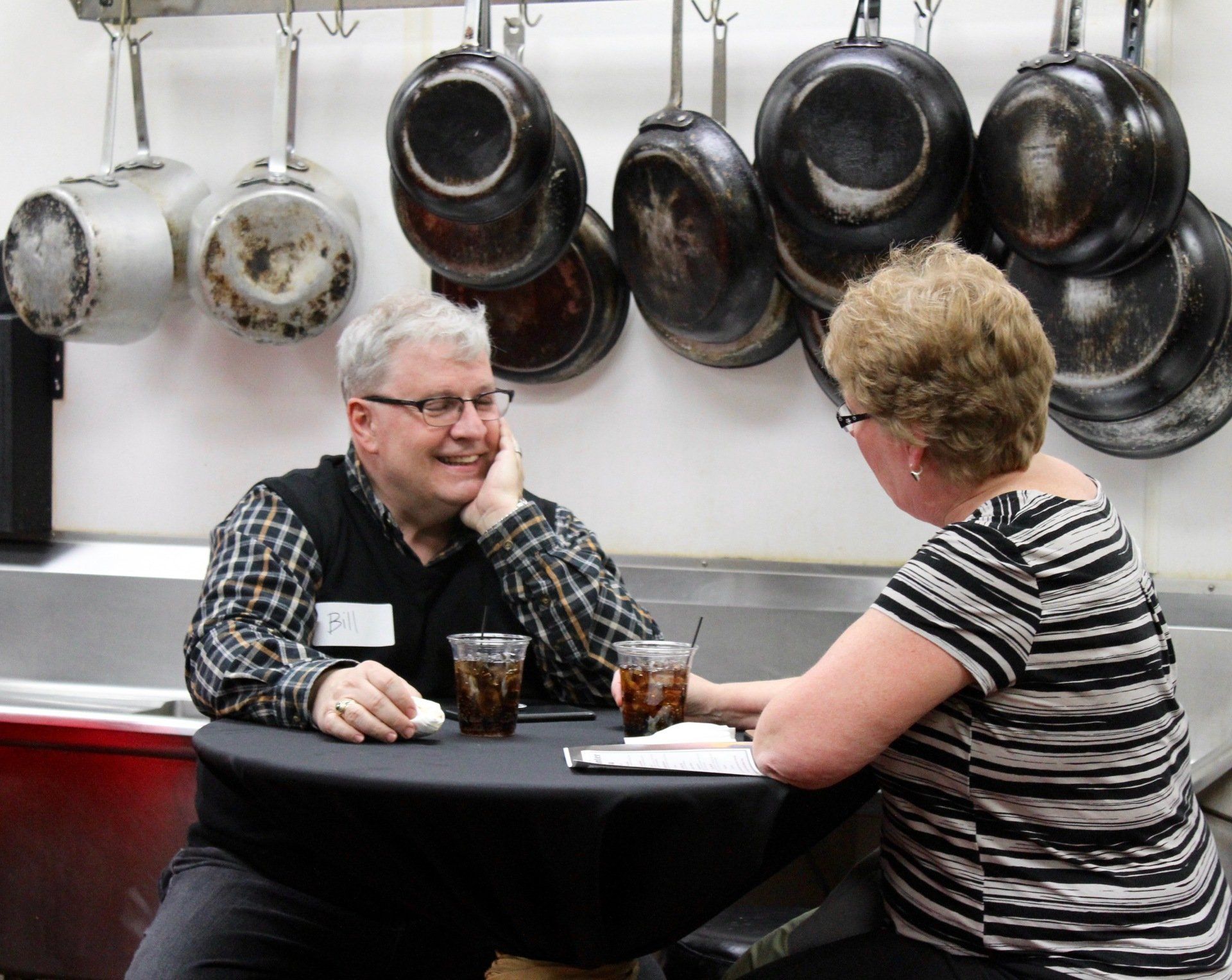 A man and a woman are sitting at a table in front of a wall of pots and pans