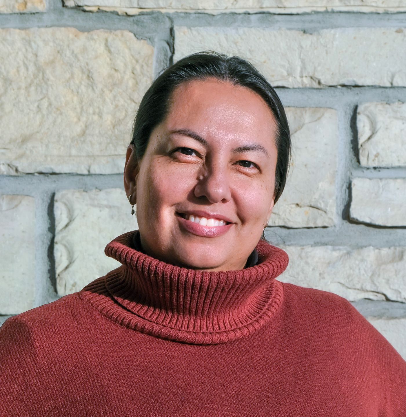 A woman wearing a red sweater is smiling in front of a brick wall