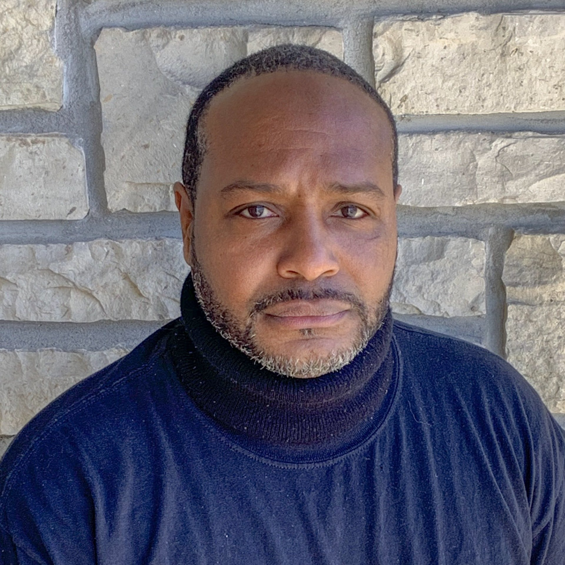 A man with a beard wearing a black turtleneck is standing in front of a brick wall.