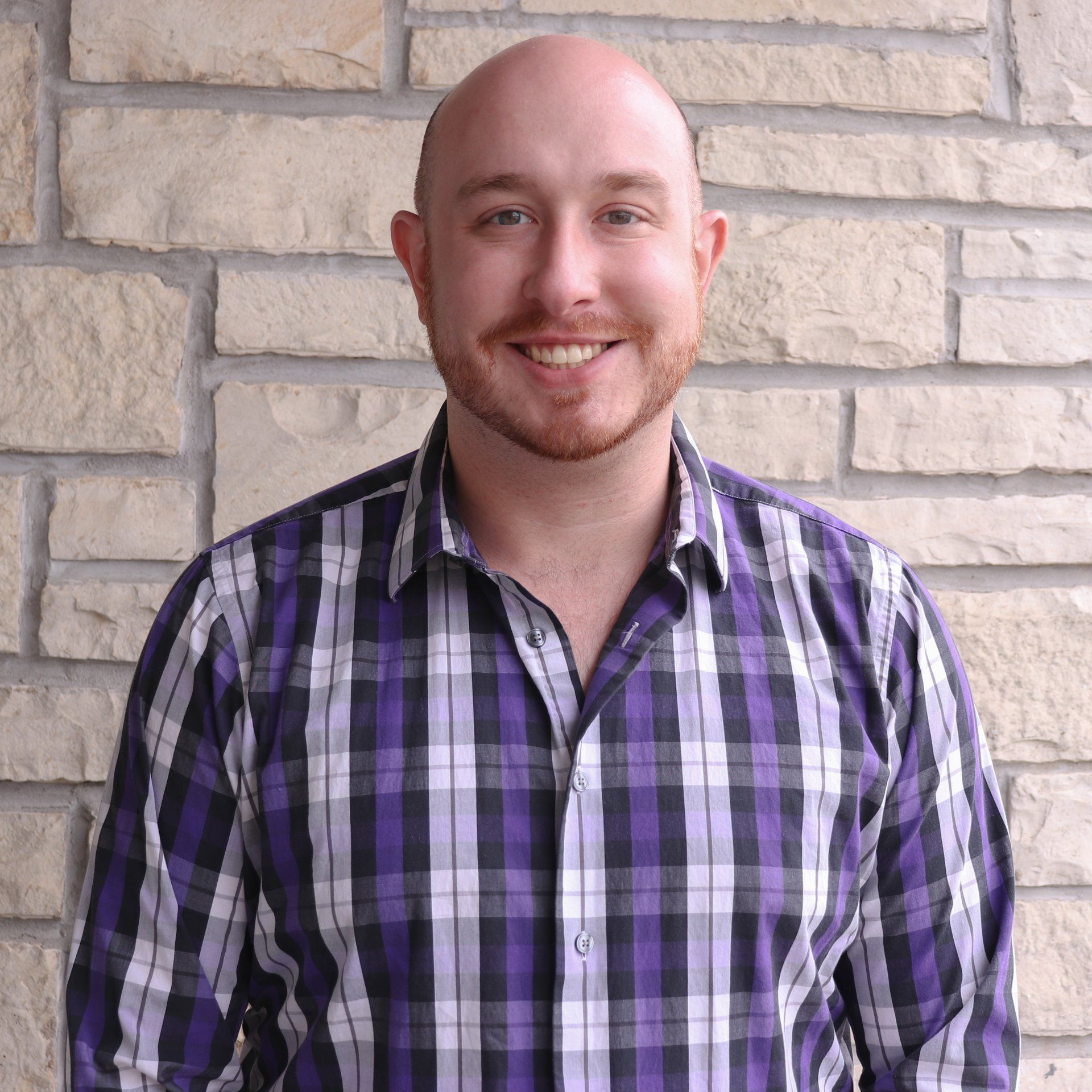 A bald man wearing a purple plaid shirt is smiling in front of a brick wall