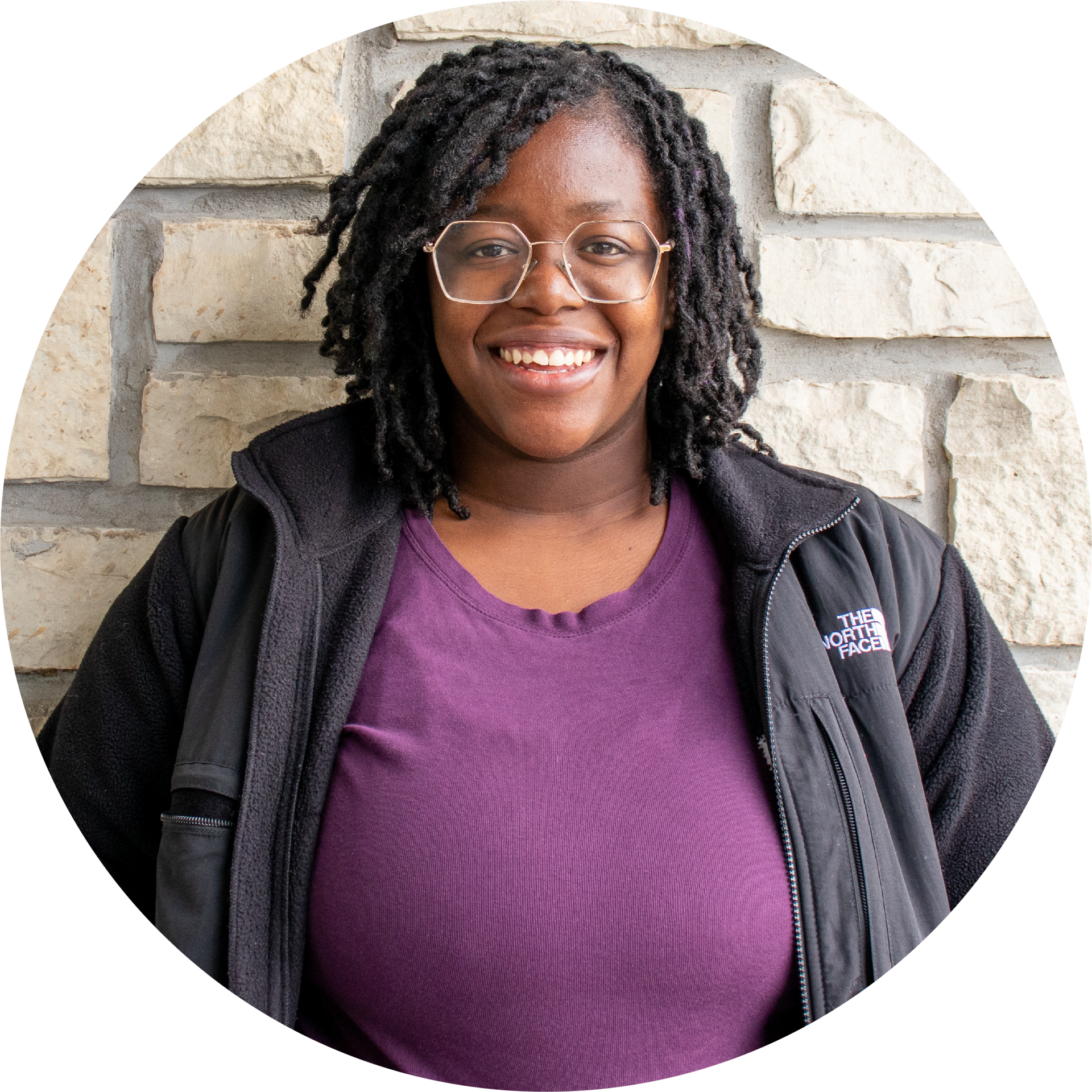 A woman wearing glasses and a purple shirt is smiling in front of a brick wall.