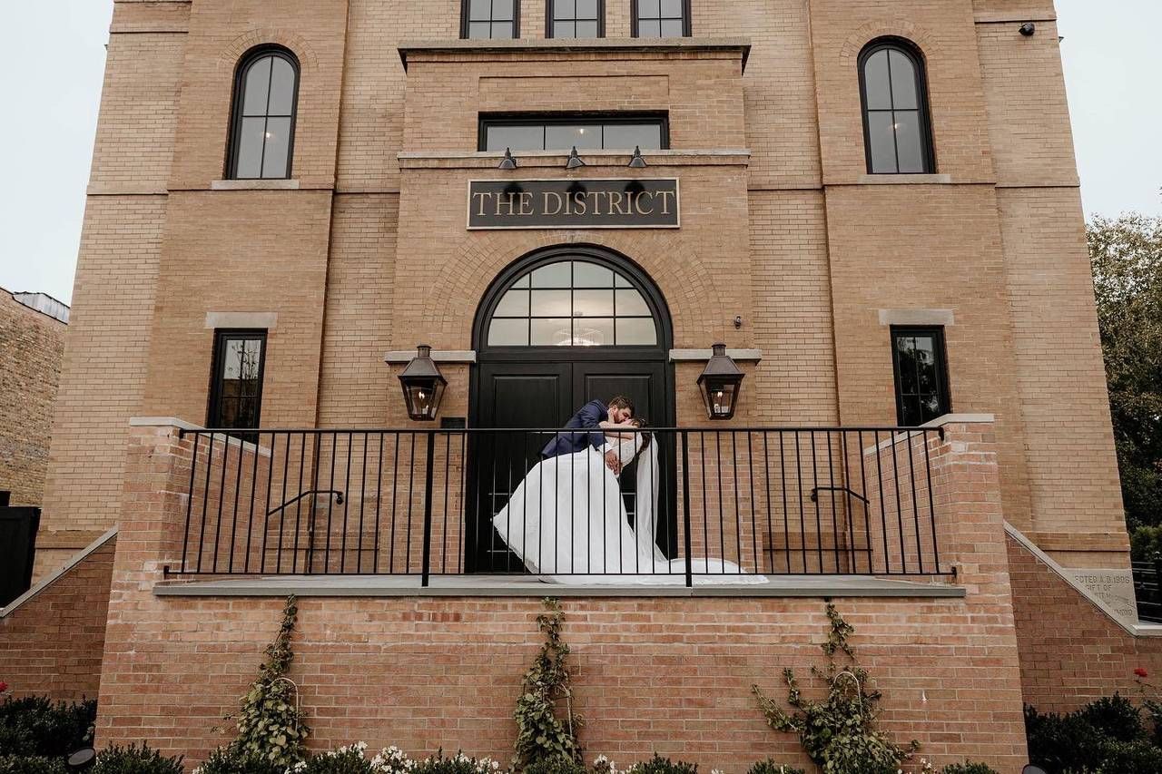 A bride and groom are kissing on a balcony in front of a large brick building.