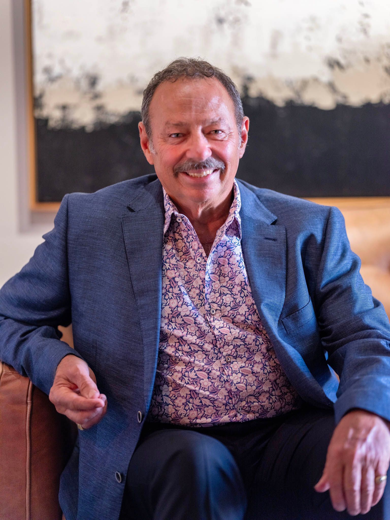 Man smiling in blue blazer and patterned shirt, seated in a room with abstract art.