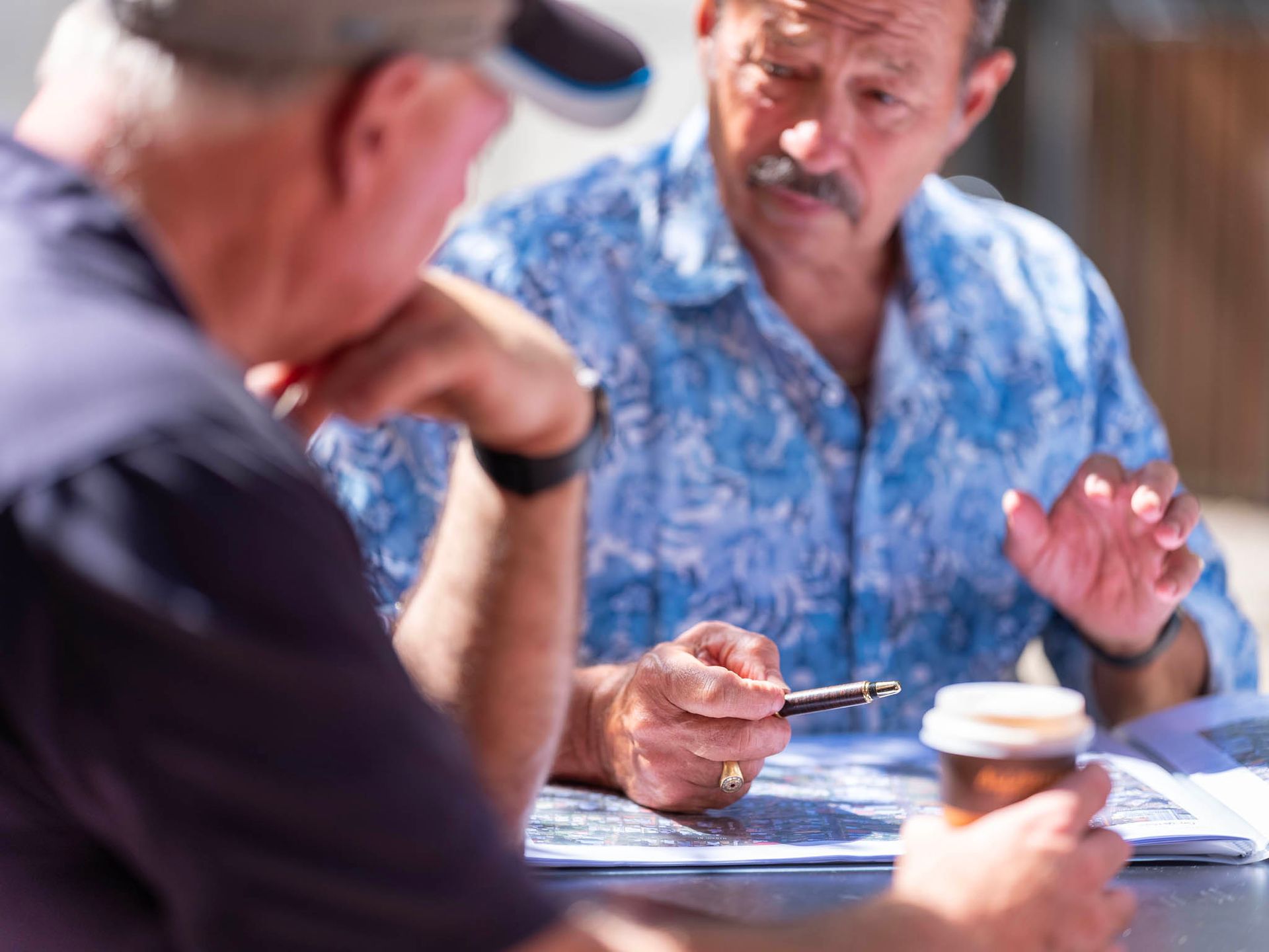 Two men seated at a table, discussing paperwork. One points with a pen, the other listens.