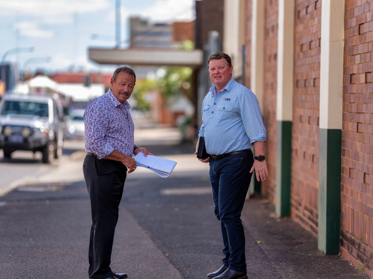 Two men stand on a sidewalk near a brick building, one holding papers. A car drives on the street.
