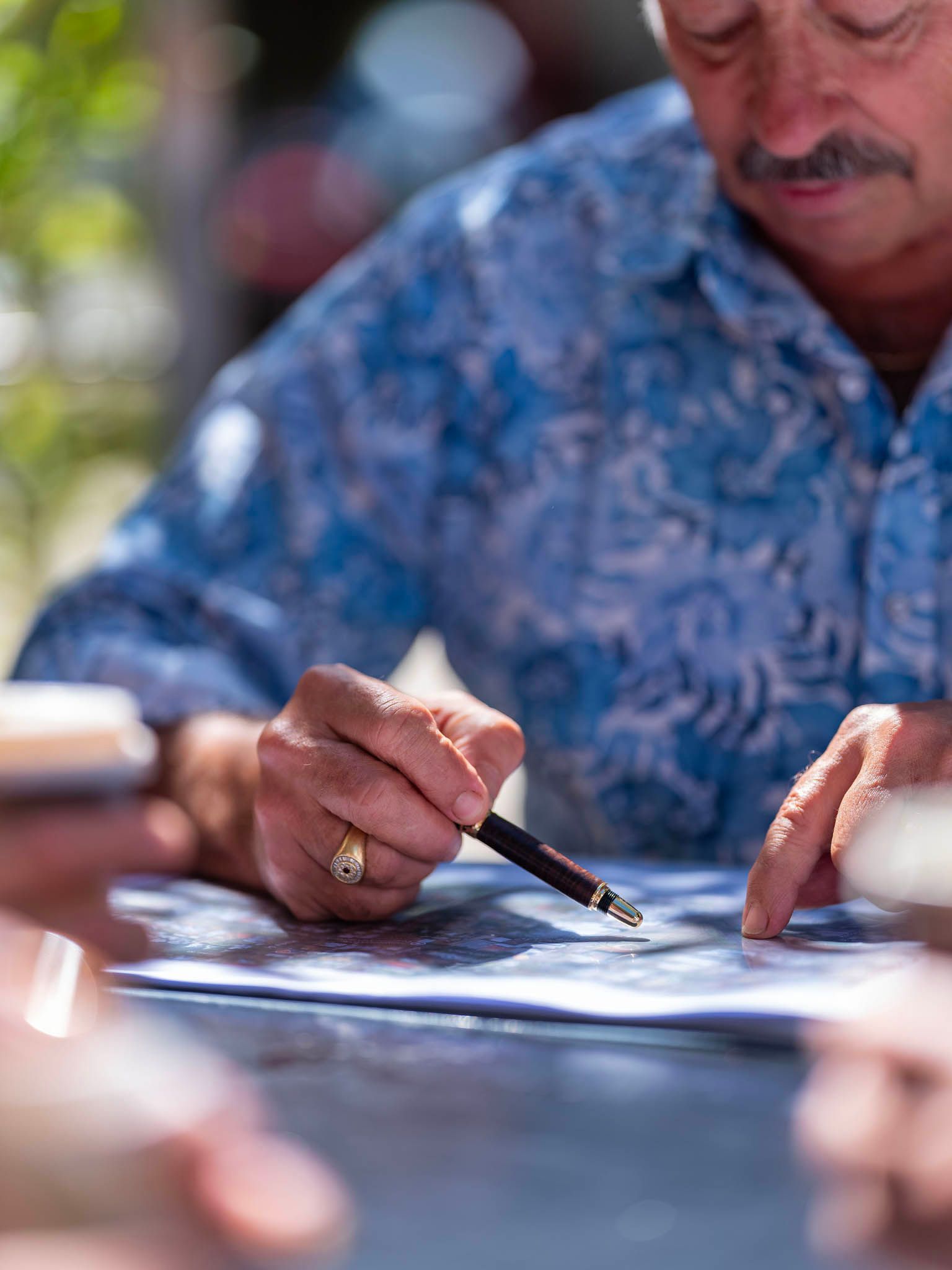 Man pointing at a map with a pen. He wears a blue floral shirt, outdoors.