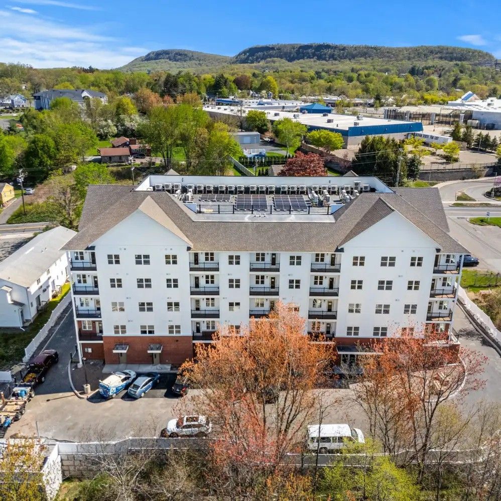 Aerial view of a white multi-story building with a brown roof and balconies, surrounded by parking and trees.