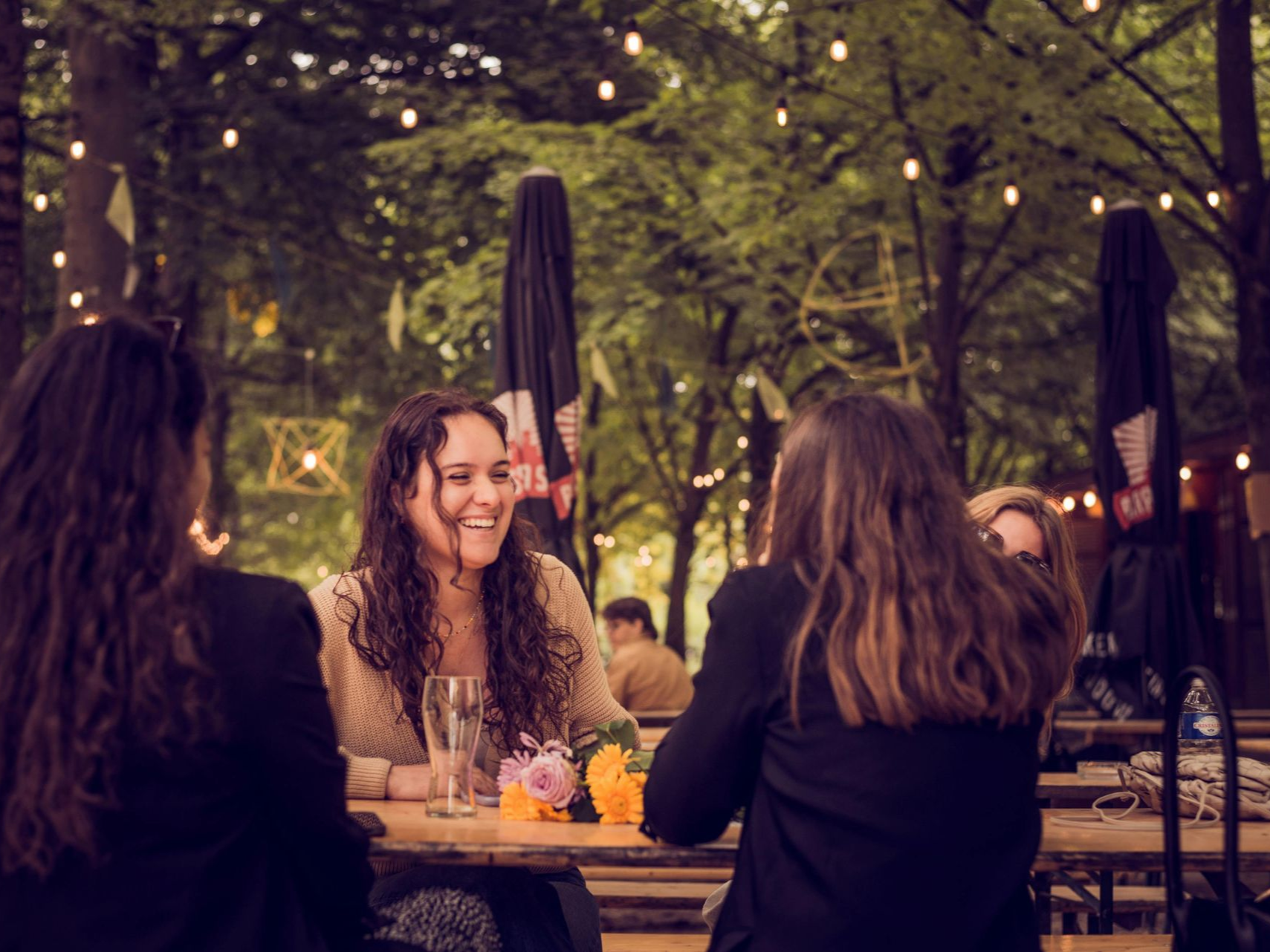 Women laughing, seated at a table outdoors, with trees and string lights.