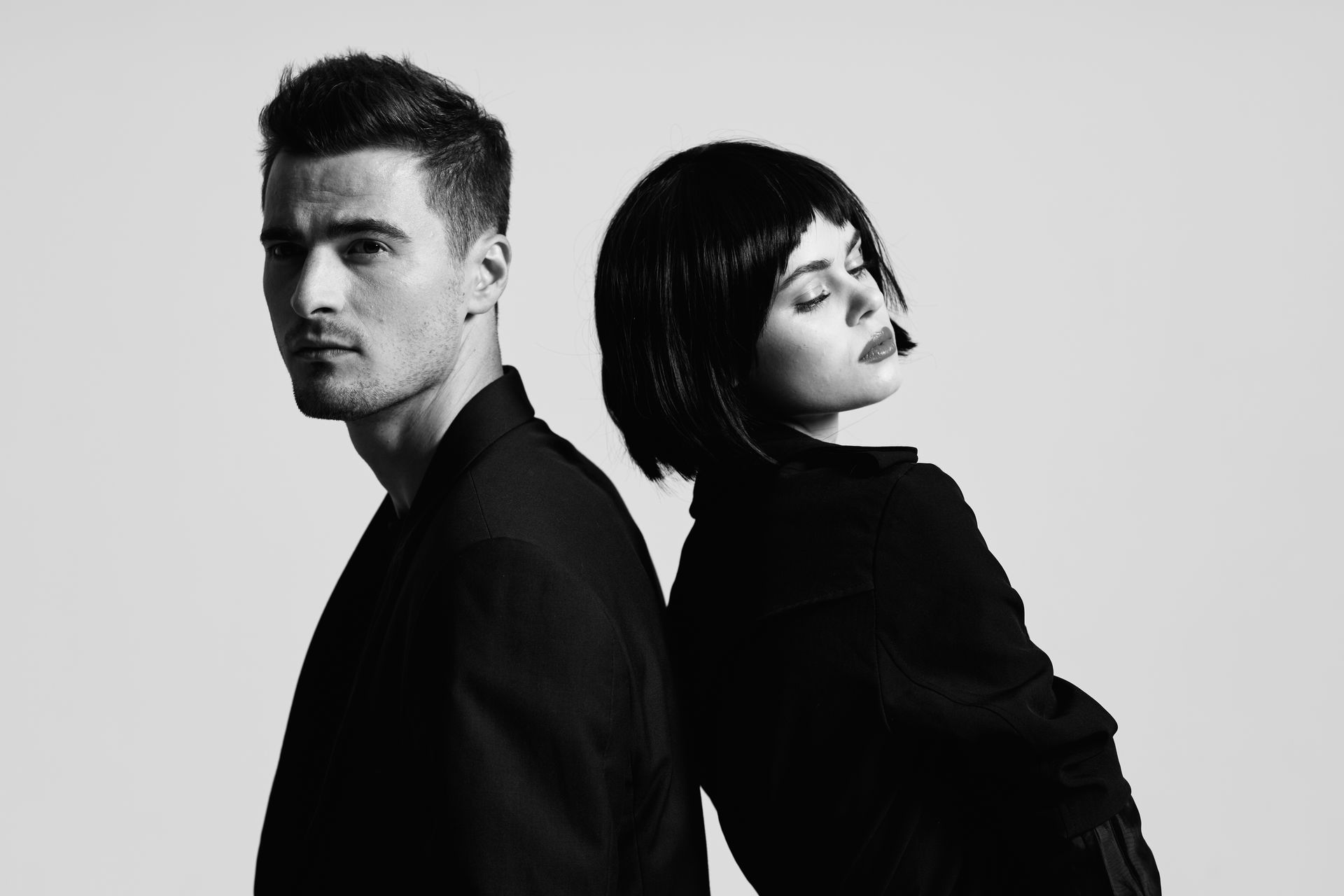 Man and woman standing back-to-back, wearing dark clothing. Woman has short dark hair, eyes closed. Black and white.