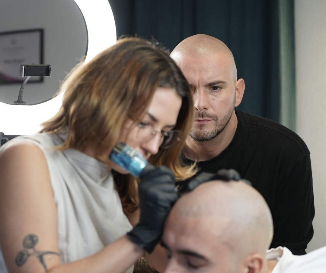 Woman tattooing a man's scalp, another man watches. Indoors, bright ring light.