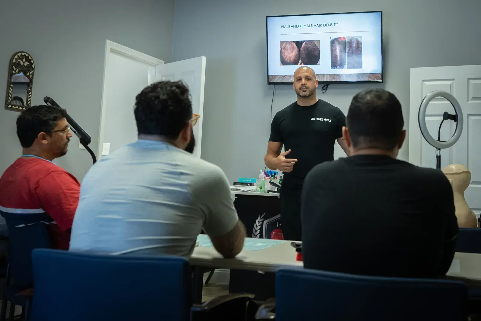A bald man presents to a group. They are sitting around a table in a room, with a presentation on a screen.