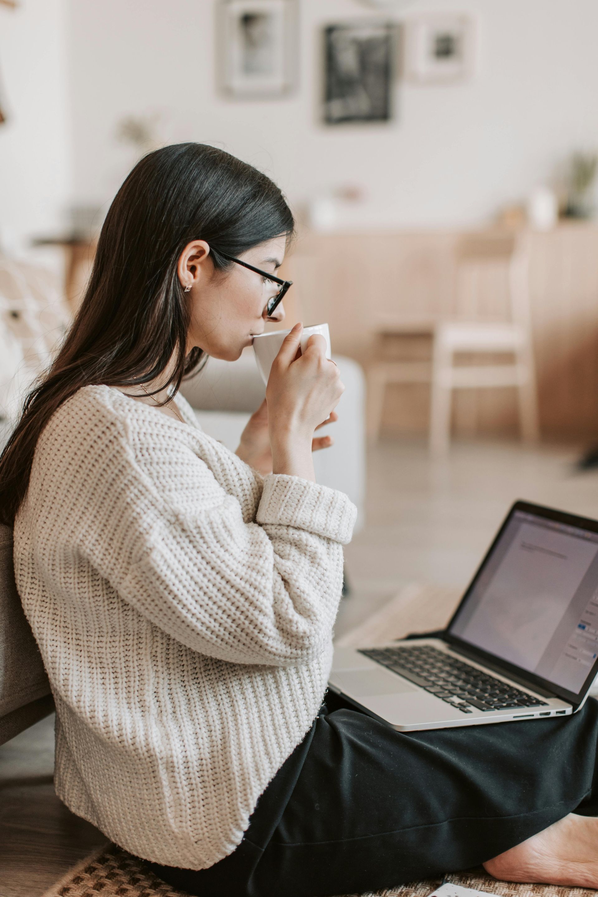 Woman sitting on floor, drinking from mug while using laptop.