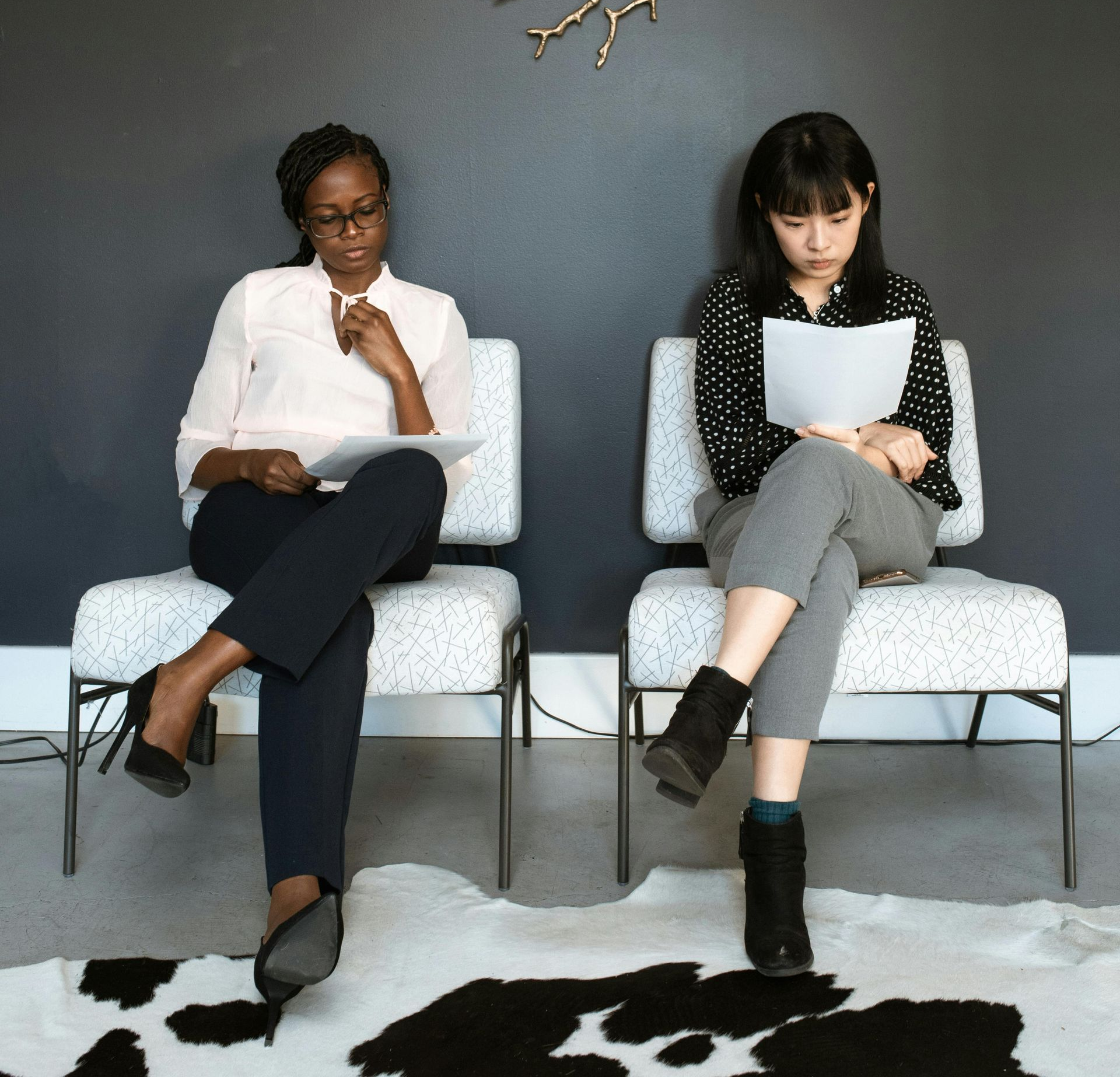 Two women in a waiting room, each holding papers. One wears glasses and a pink shirt. The other wears a black top.