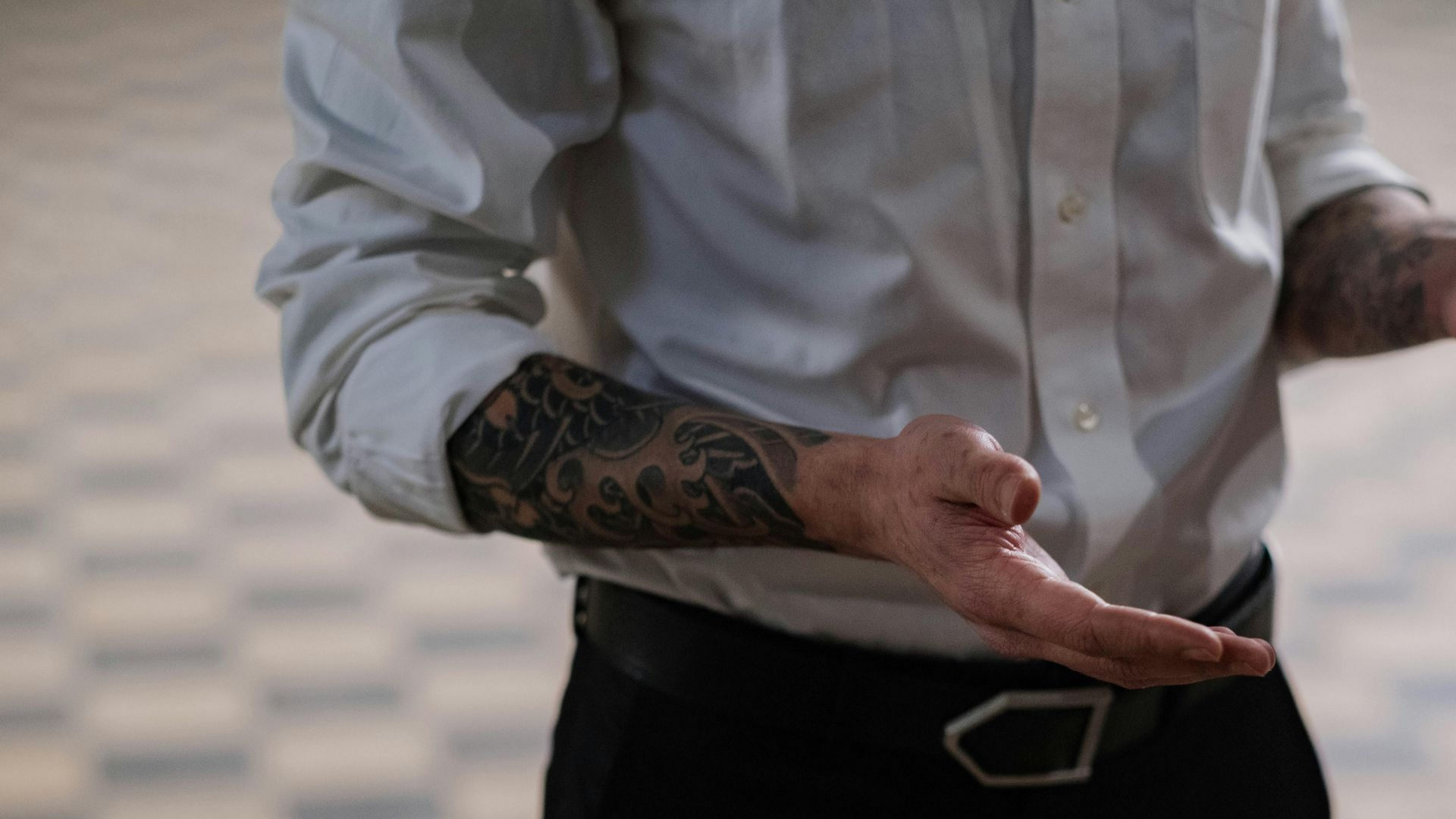 Man with sleeve tattoos, gesturing with hands. White shirt, black pants, belt, checkered floor.