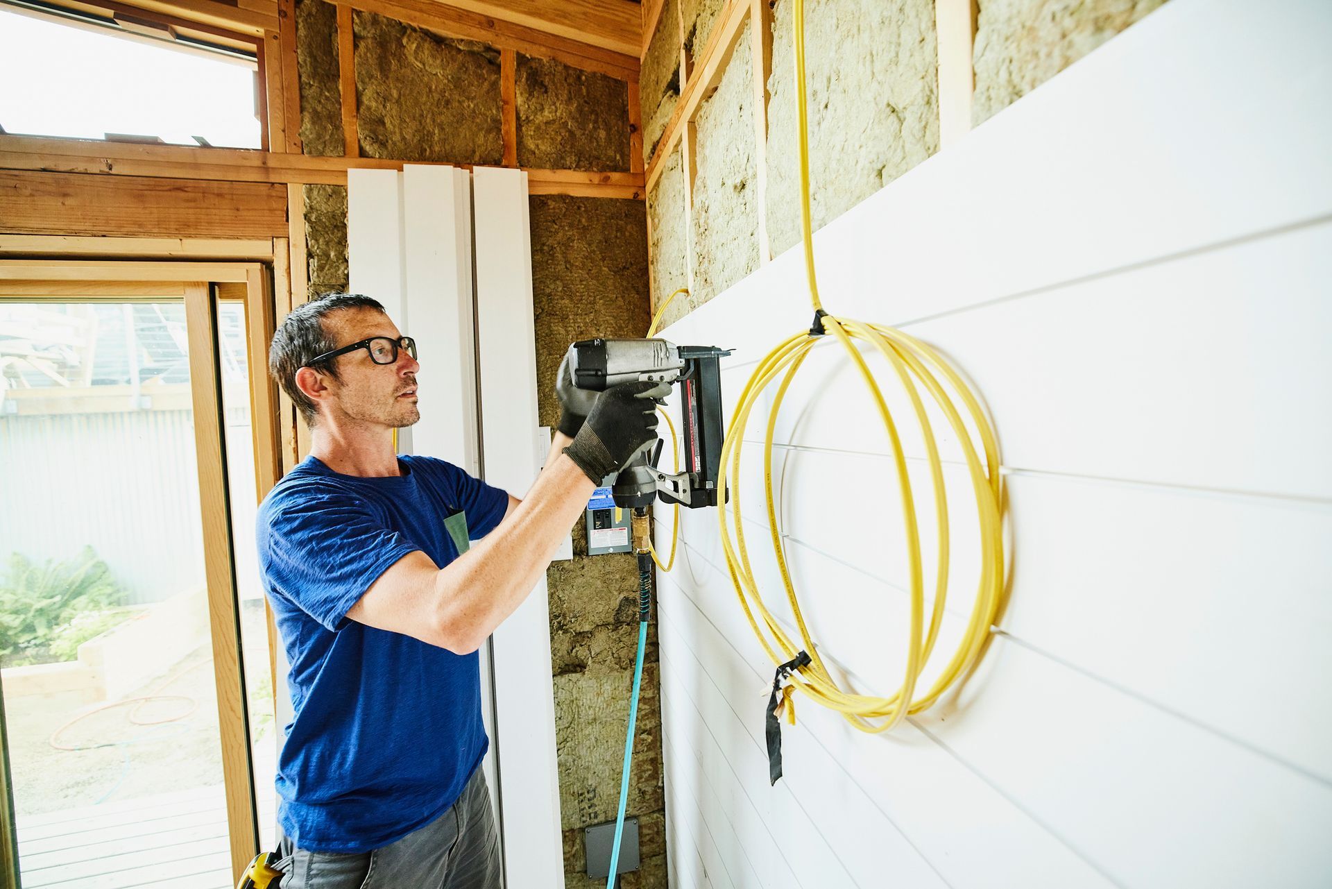 A man using a nail gun to install siding as part of a home remodeling project outdoors.