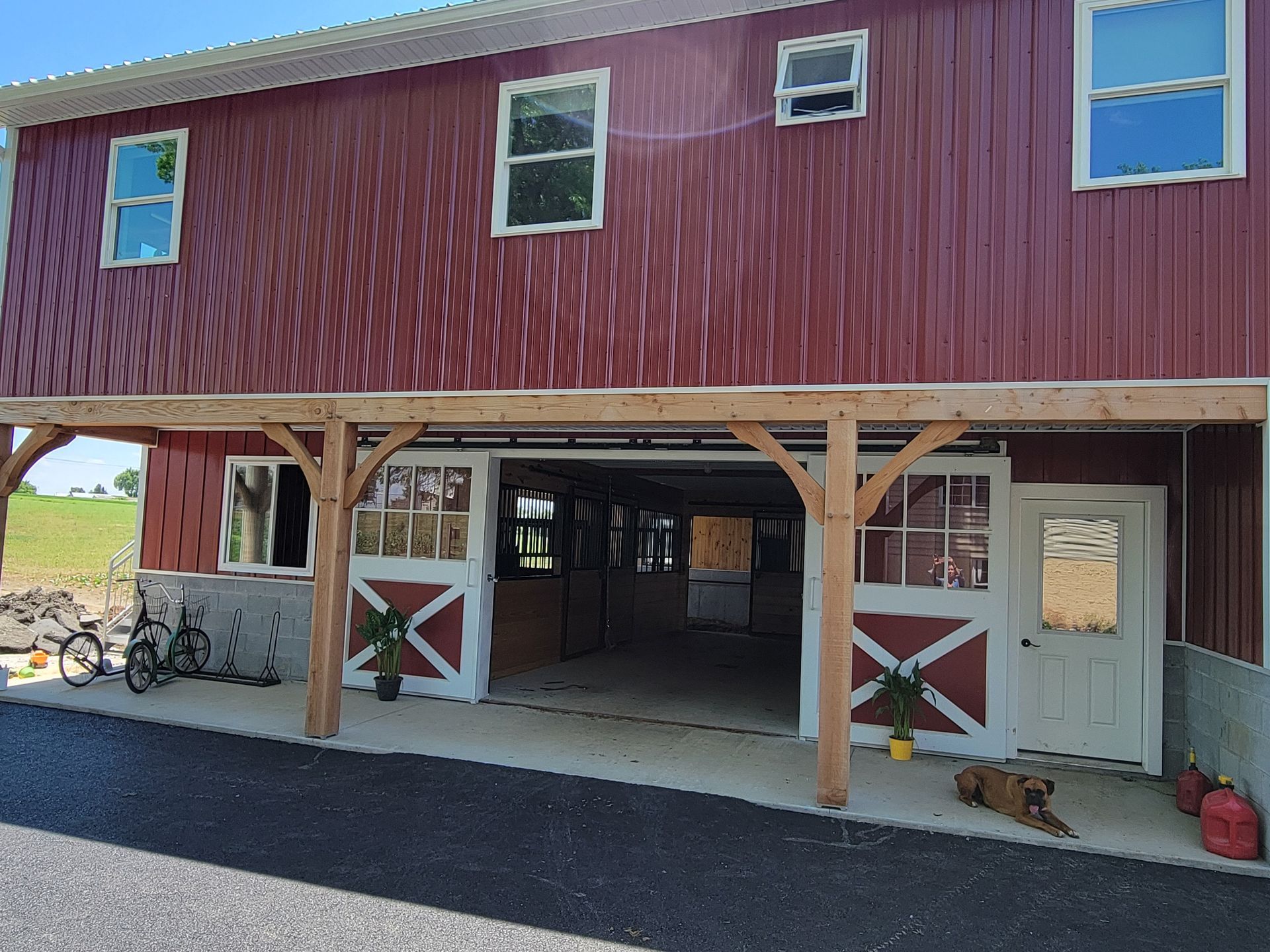Red barn with open doors, concrete base, and white trim. Bike, dog, and potted plants are visible.