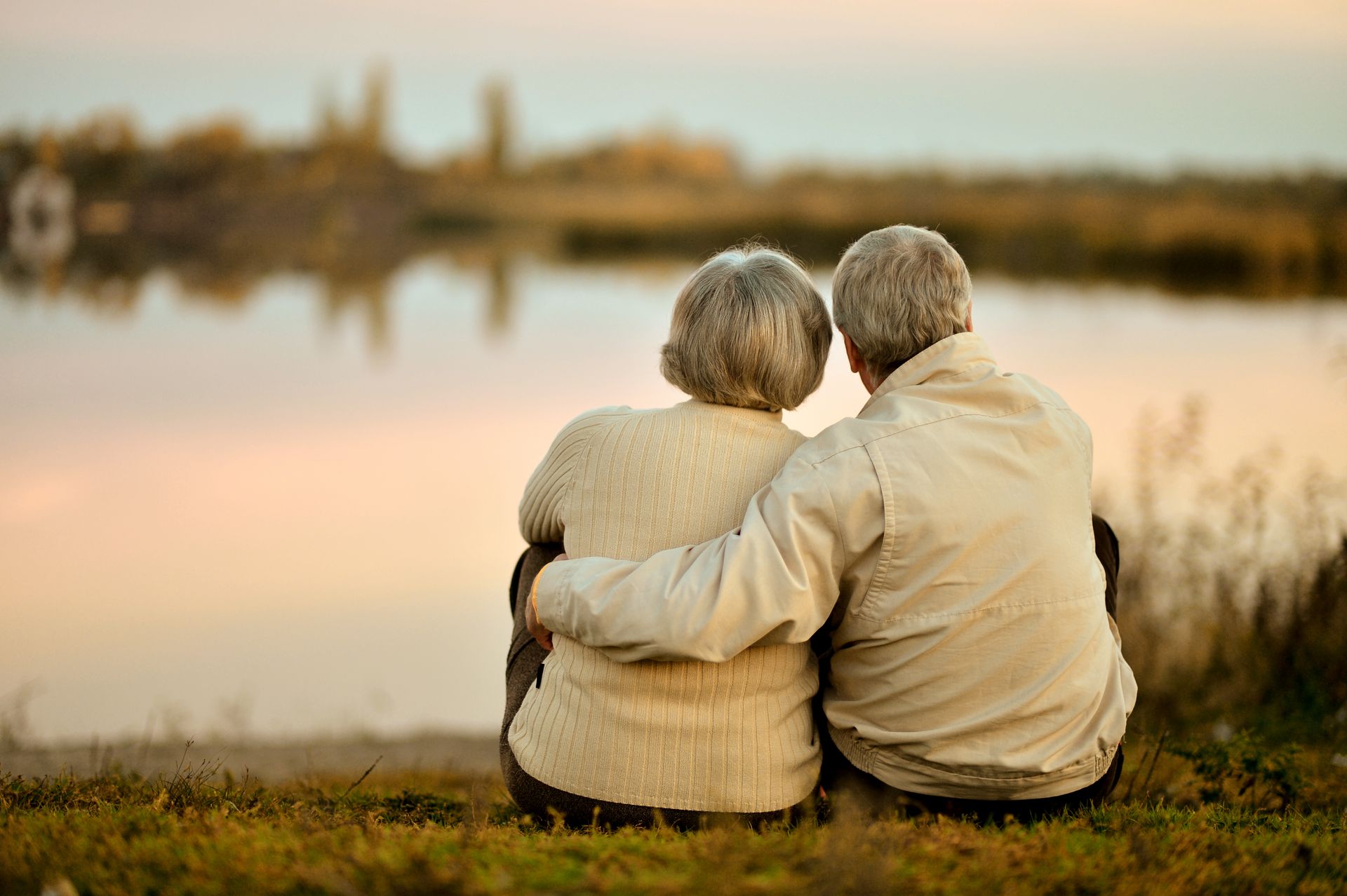 An elderly couple is sitting on the grass looking at a lake.