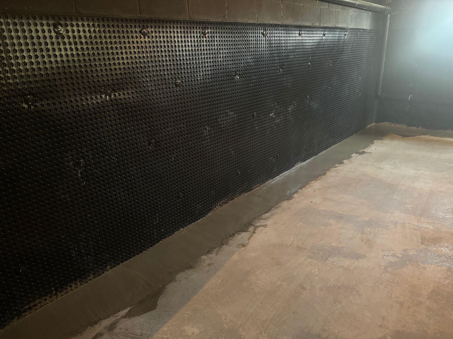 A basement wall featuring black, dimpled waterproof drainage membrane installed above a fresh concrete floor seal.