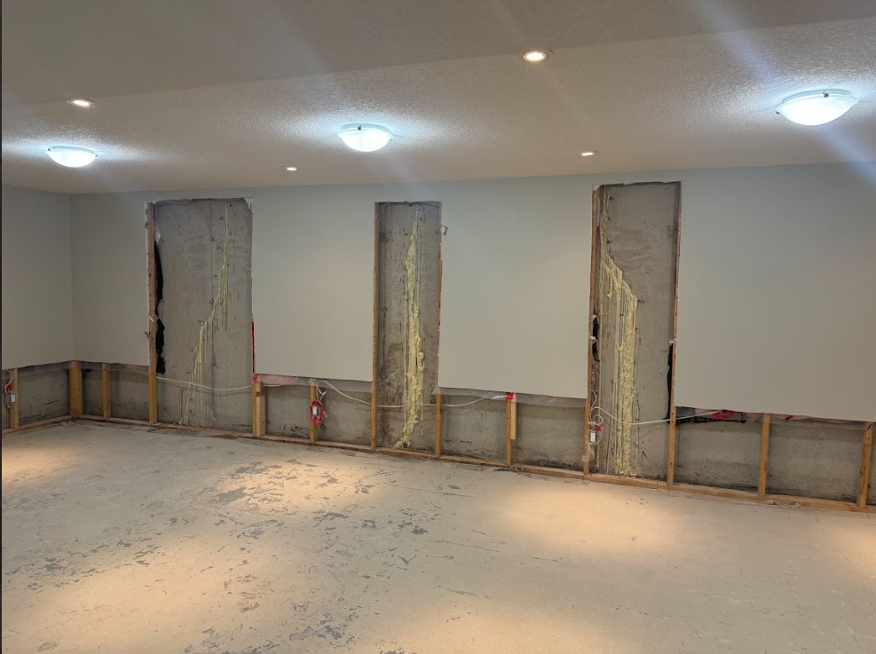 Basement under construction; exposed concrete pillars, wooden framing, bare concrete floor, three ceiling lights.