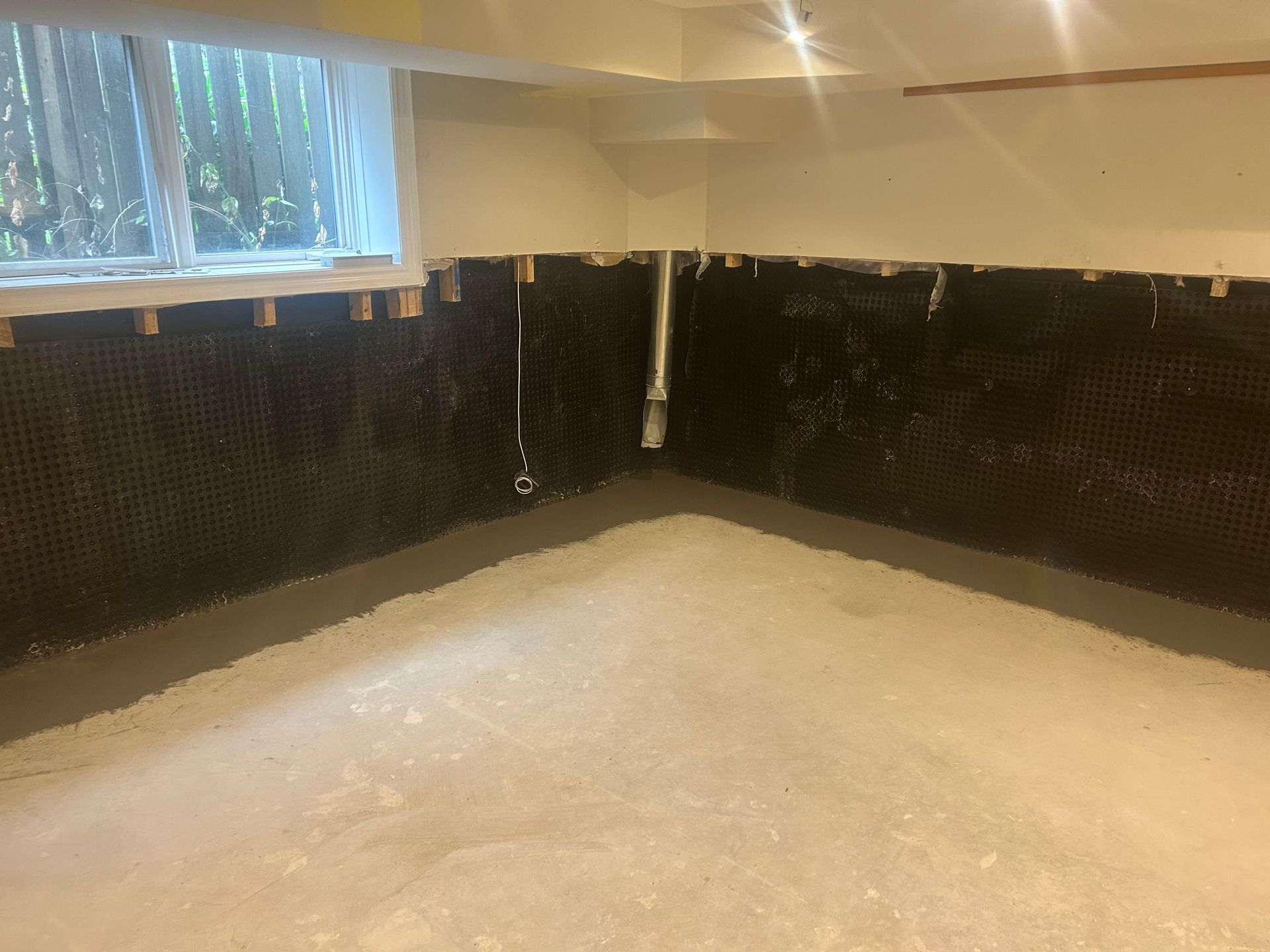 A basement corner with unfinished concrete floors and walls lined with black dimpled waterproofing membrane.
