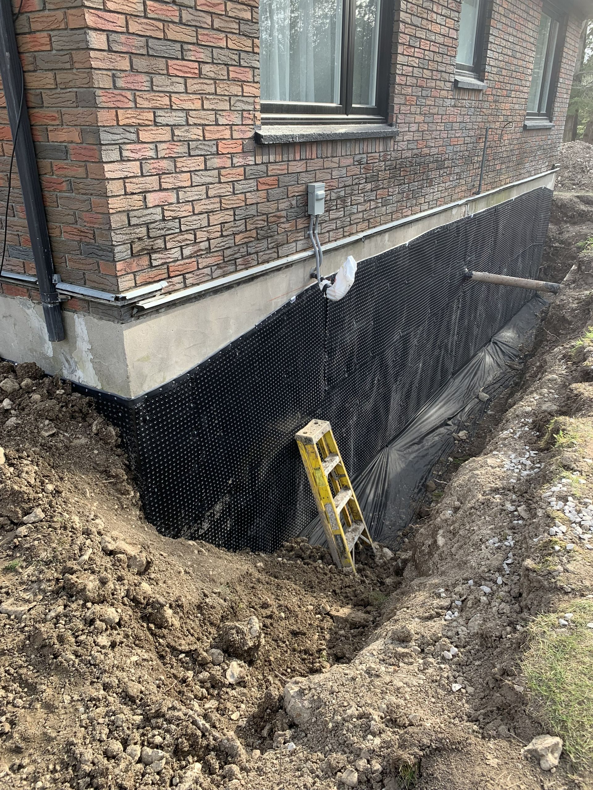 Foundation waterproofing in progress: black dimpled membrane against brick wall, trench with soil, level leaning on wall.