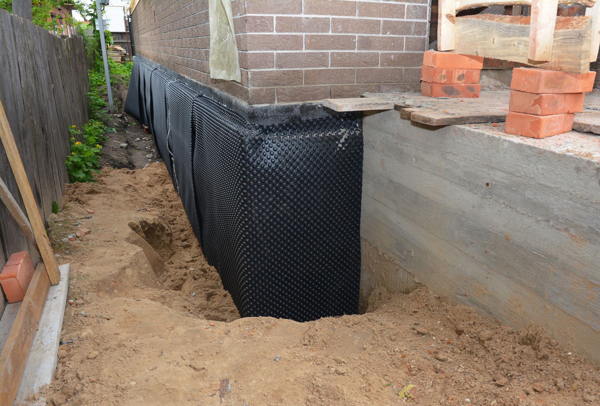 A black dimpled drainage membrane is installed against a concrete foundation wall in an excavated residential side yard.