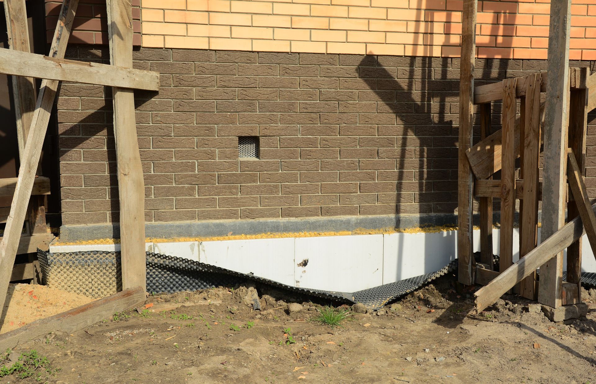 A building foundation showing exposed white insulation, a protective black mesh, and brick masonry under construction.