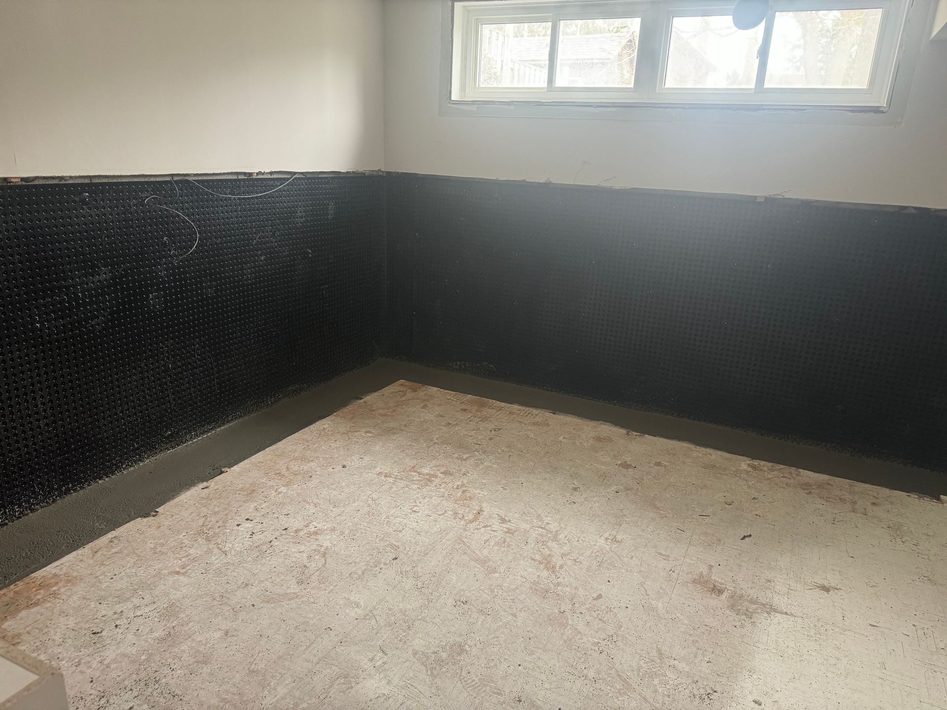 A basement corner with black dimpled waterproofing on the walls, window, and concrete floor.